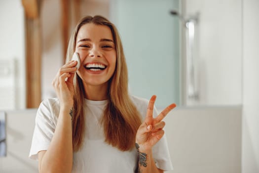 Woman wiping her face with cotton pad in the bathroom and showing sign peace. Skin care concept