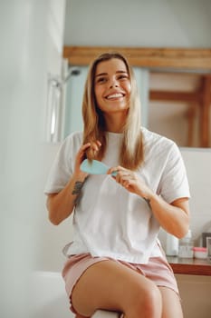 Smiling young woman brushing her hair with comb while standing in bathroom near mirror