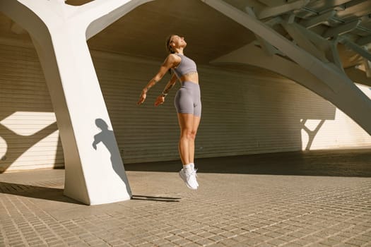 Female athlete in sportswear leaping in air with wall backdrop outdoors. Healthy lifestyle concept