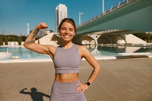 Female model in sportswear showing biceps standing outdoors and looks camera with smile