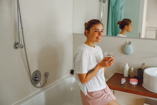 Young beautiful woman drinking glass of water in the bathroom at home. Beauty treatment