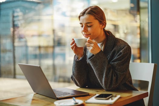 Smiling female entrepreneur is drinking coffee in coworking while working on laptop near window