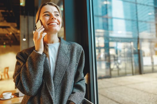 Smiling businesswoman is talking phone with colleagues while standing in cafe near window