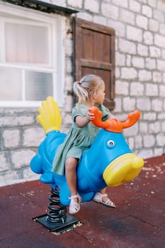 Little girl swings on a spring swing in the shape of a deer in the yard of the house looking back. High quality photo