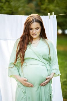 White pretty pregnant woman stands putting hands on big belly. Green trees, park,white sheet on background. Young healthy female wears dress. Baby shower,child delivery, preparation concept. Vertical
