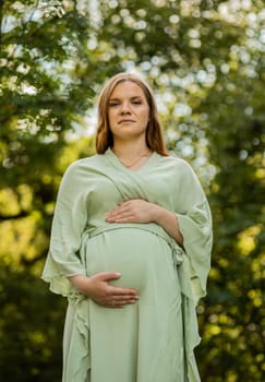Young pregnant woman puts hands on big belly, green trees,blue sky on background. Female wears dress. Magic happy pregnancy. Labor,emotional connection with baby. Vertical plane