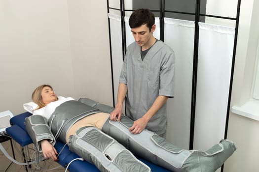 Male therapist prepares pressure therapy procedure, massaging suit for female client in spa salon. Pressotherapy, lymphatic drainage massage. Horizontal plane, copy space
