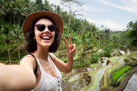 Young woman traveler wearing hat and sunglasses taking selfie in Tegalalng rice terrace, Bali. Female taking picture in summer vacation. Travel, freedom and happiness concept.