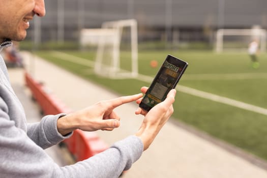 Portrait of handsome smiling bearded man being happy to win at bookmaker's mobile application after betting at favourite football team, stadium on the background.