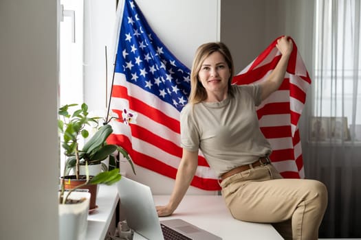 Portrait of happy young woman with USA flag learning American English online from home using laptop. Smiling female student sitting at table and taking online educational course in foreign languages. High quality photo