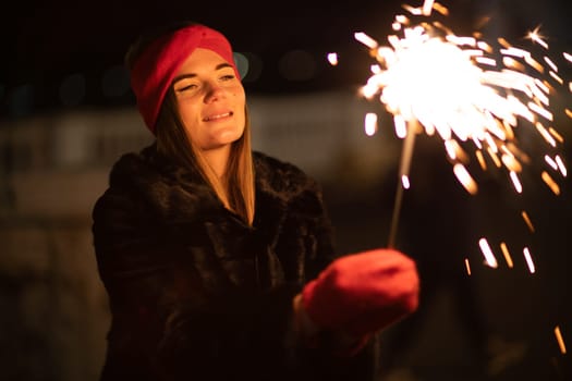 Woman holding sparkler night while celebrating Christmas outside. Dressed in a fur coat and a red headband. Blurred christmas decorations in the background. Selective focus.