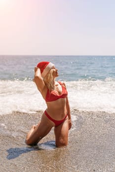 Female beach Santa hat wave coast. beach relaxation seaside. A woman in a red swimsuit enjoying her time on the beach, sits on the sand and being covered by a wave