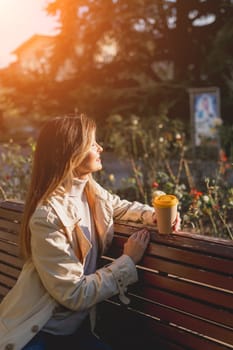 Woman drinks from cup on wooden bench. She is wearing a white shirt enjoying her beverage. The bench is located in a park setting, with trees in the background