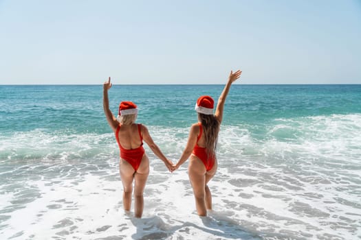 Women Santa hats ocean play. Seaside, beach daytime, enjoying beach fun. Two women in red swimsuits and Santa hats are enjoying themselves in the ocean waves and raising their hands up