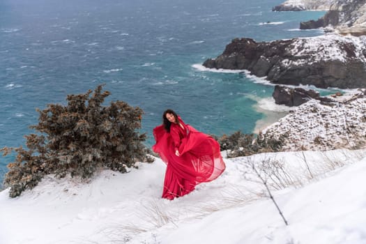 Woman red dress snow sea. Happy woman in a red dress in the snowy mountains by the emerald sea. The wind blows her clothes, posing against sea and snow background