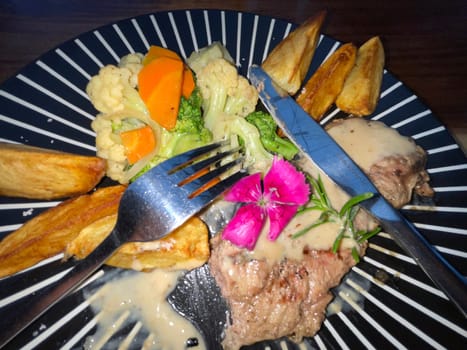 Juicy beef steak with salad and potato wedges along with pepper corn sauce served on black and white stripe on a wooden stone table. Top view