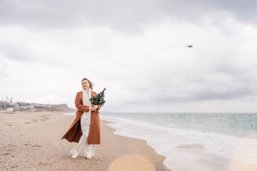 Blond woman Christmas sea. Christmas portrait of a happy woman walking along the beach and holding a Christmas tree in her hands. She is wearing a brown coat and a white suit