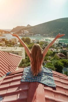 Woman sits on rooftop with outstretched arms, enjoys town view and sea mountains. Peaceful rooftop relaxation. Below her, there is a town with several boats visible in the water.