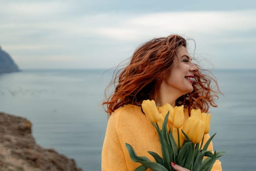 Portrait of a happy woman with hair flying in the wind against the backdrop of mountains and sea. Holding a bouquet of yellow tulips in her hands, wearing a yellow sweater.