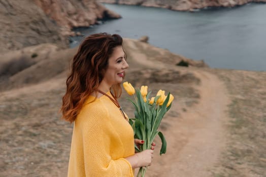 Portrait of a happy woman with long hair against a background of mountains and sea. Holding a bouquet of yellow tulips in her hands, wearing a yellow sweater.