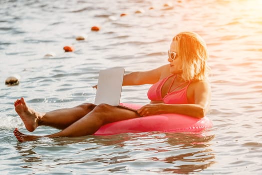 Woman works on laptop in sea. Freelancer, young blond woman in sunglases floating on an inflatable big pink donut with a laptop in the sea at sunset. Freelance, travel and holidays concept