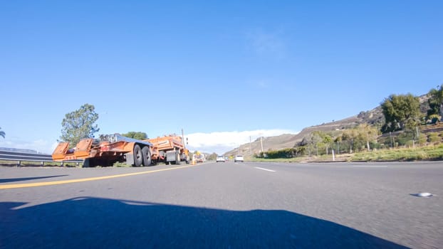 On a crisp winter day, a car cruises along the iconic Highway 1 near San Luis Obispo, California. The surrounding landscape is brownish and subdued, with rolling hills and patches of coastal vegetation flanking the winding road.