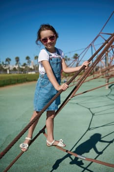 Little child girl climbing on the climbing net in the urban playground. Full length portrait of cute kid in casual denim playing on the city playground climbing and jumping on the trampoline
