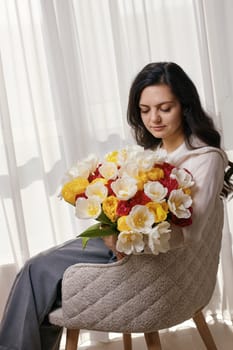 Beautiful woman with spring flowers tulips in hands sitting on chair near window. Women's Day