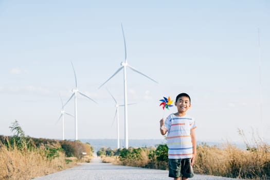 Cheerful child engaging with wind energy holding a pinwheel near turbines captures spirit of playful innovation. Showcases clean electricity amidst a countryside windmill farm under a serene blue sky.