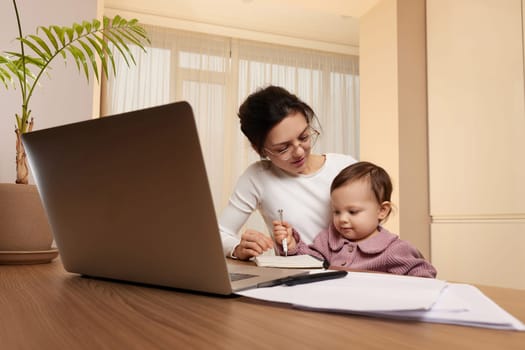 positive businesswoman in glasses working on laptop at home with her little child girl. Stay at home