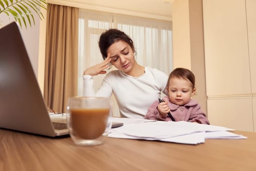 Stressed frustrated woman working on laptop at home with her little baby girl. Child makes noise and disturbs mother at work.