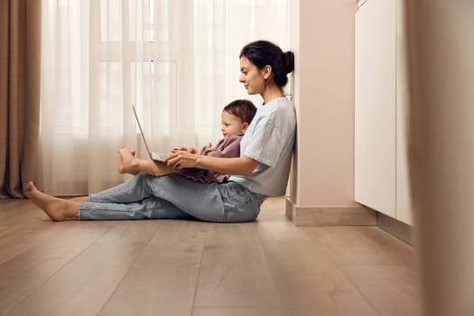 beautiful casual woman sitting on the floor and working on laptop with her little child girl at home