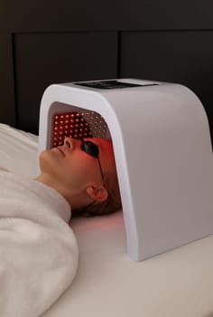 Cosmetic Red Led Light Face Mask. Woman In Her 30s Lying Under Facial Regenerative Treatment Mask On Bed In Bedroom. Home Skin Care Procedure. Acne, Spot Cure. Beauty Photon Therapy. Vertical Plane.