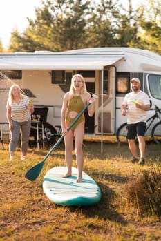 the family is resting next to their mobile home. My daughter is standing with a paddle on a sup board, and her parents pour water on her.