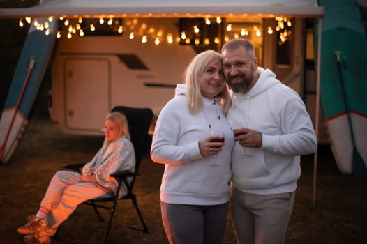 A married couple with glasses of wine stands against the background of a motorhome and rests together by the campfire. Evening family vacation.