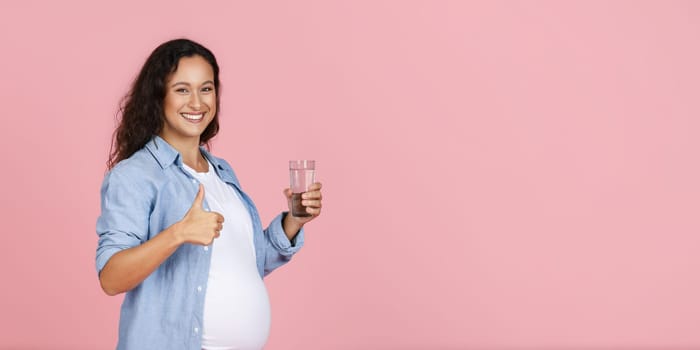 Hydration during pregnancy, healthy lifestyle for expectants. Positive pregnant woman holding glass of water and showing thumb up at camera, pink studio background, copy space, web-banner