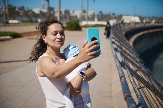 Young mother carrying her newborn baby sleeping in her arms, taking a selfie on her modern smart mobile phone, sharing her maternity lifestyle on social media, while waling on the city promenade