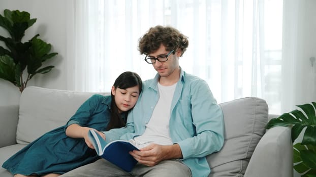 Happy daughter lean on father shoulder while listening story telling. Young asian schoolgirl looking ar dad while reading story book together and sitting at sofa. Family recreation concept. Pedagogy.