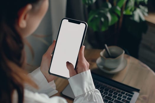 A woman is sitting at a table, gesturing with her thumb on a gadget, the cell phone with a white screen. Her fingers tapping on the font space bar, her thigh resting on a plant event