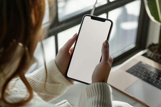 The woman is holding a communication device with a white screen in her hands, using her thumb to interact with the gadget. She is sitting at a desk, with her eyewear on the table and making a gesture