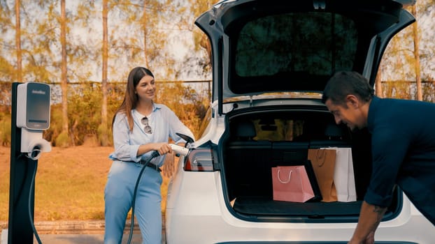 Couple recharge their EV car with electric battery charging station at rest stop during their road trip travel during autumnal season to national park. Relaxing holiday with eco friendly car. Exalt