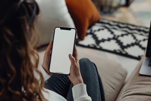 A woman is seated on a couch, displaying a gesture of holding a cell phone with a blank white screen. Her thumb rests on the device as she exudes comfort and casual fashion