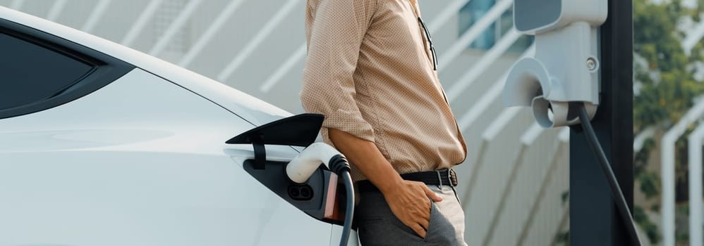 Young man recharge electric car's battery from charging station in city commercial parking lot. Rechargeable EV car for sustainable environmental friendly urban travel. Panorama Expedient