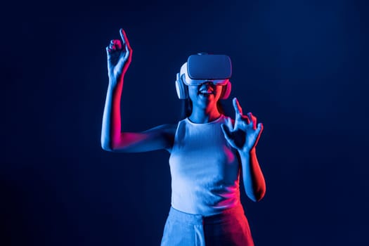 Smart female standing in cyberpunk neon light wear VR headset connecting metaverse, futuristic cyberspace community technology. Elegant woman use finger point generated virtual object. Hallucination.