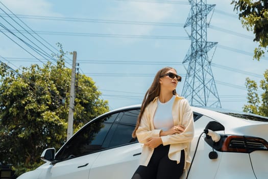 Young woman recharge EV car battery at charging station connected to power grid tower electrical industrial facility as electrical industry for eco friendly vehicle utilization. Expedient
