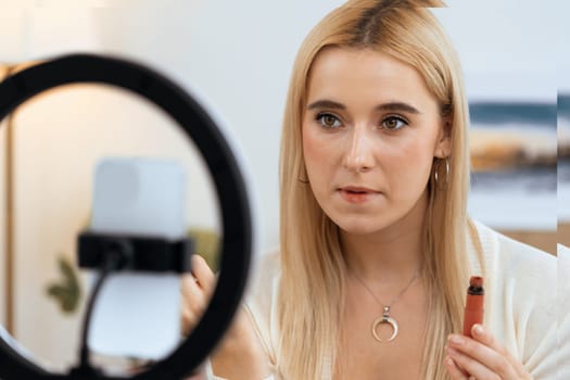 Young woman making beauty and cosmetic tutorial video content for social media. Beauty blogger using camera and light ring while showing how to apply liquid lipstick to audience or follower. Blithe