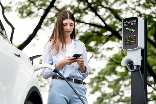 Young woman using smartphone online banking application to pay for electric car battery charging from EV charging station during vacation holiday road trip at national park or summer forest. Exalt