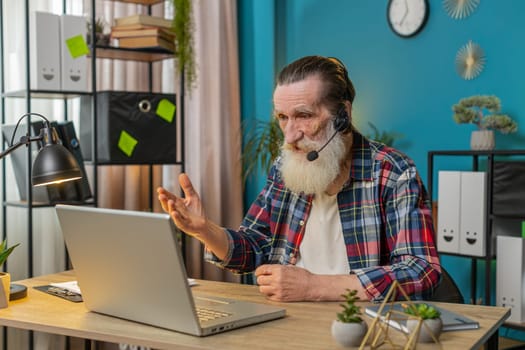 Caucasian senior businessman wearing headset, freelance worker, call center or support service operator helpline, talking with client communication support. Grandfather sitting at home office desk.