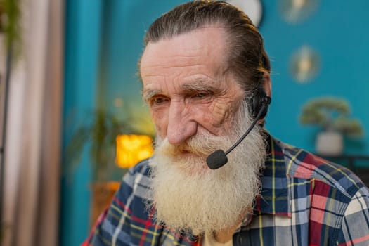 Close-up senior mature businessman wearing headset, freelance worker, call center or support service operator helpline, talking with client communication support. Grandfather sitting at home office