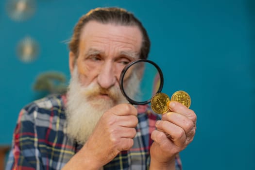Close-up face surprise senior mature man examining golden bitcoins BTC with magnifying glass. Happy elderly bearded grandfather showing gold coins sitting and looking at camera at home office.